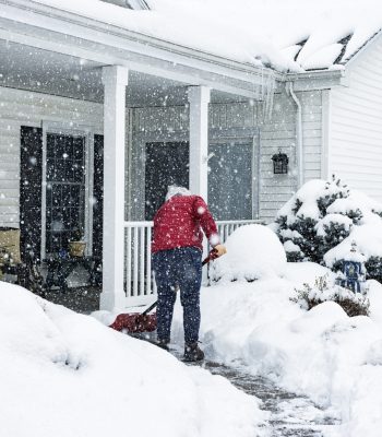 A senior adult woman wearing a warm red winter jacket coat is using a manual snow shovel to push and clear away the pedestrian walkway footpath in front of her house of deep winter snow while a western New York State February blizzard storm continues dropping more in this suburban residential district near Rochester, New York.