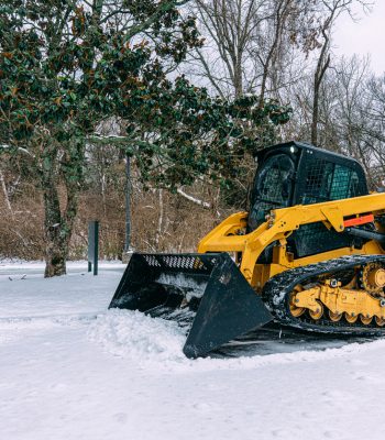 Professional Snow Removal Company or a City Worker using a Bulldozer to scrape away snow and ice from a parking lot in Midwest USA in the Winter, on an overcast day. 
They provide a clean, safe, anti-slip sidewalk and parking lot surface to protect pedestrians, tourists, customers, and local businesses.